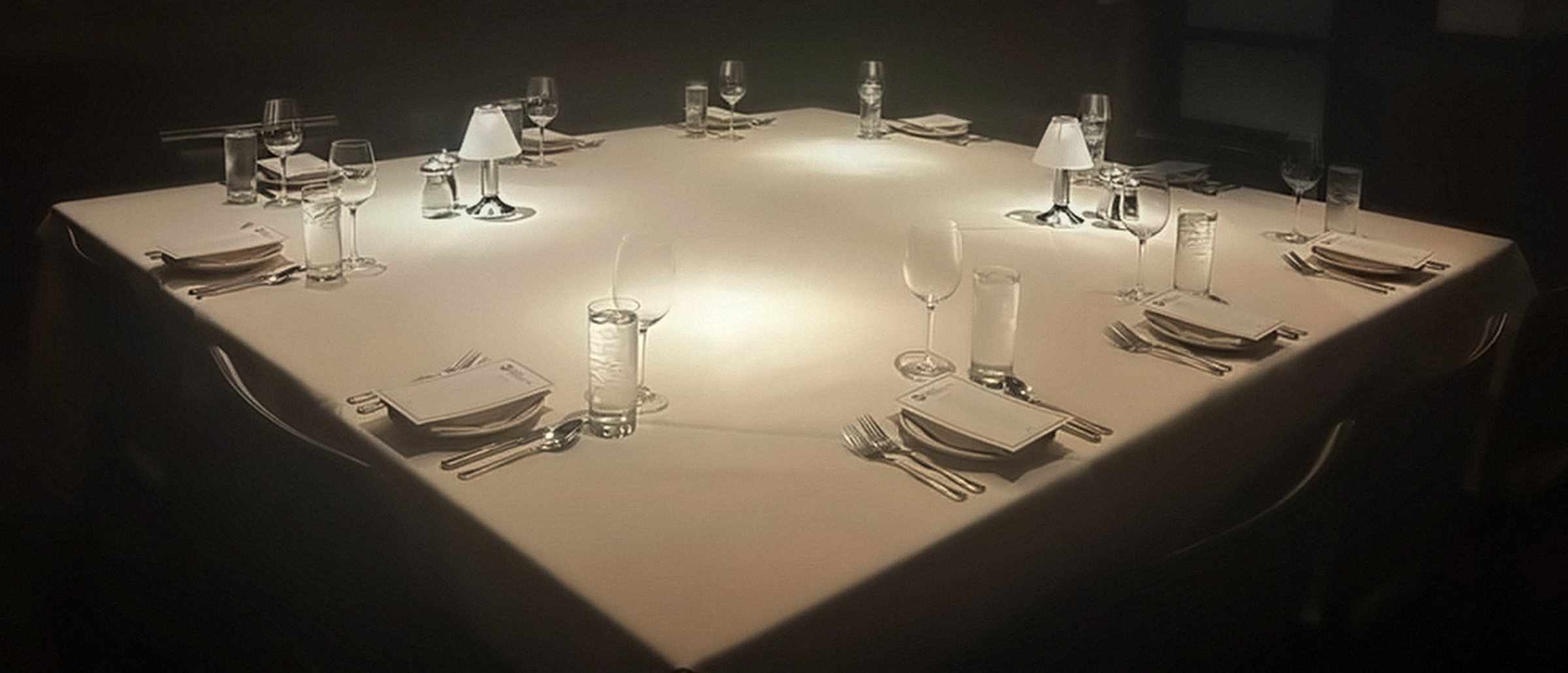 A dinner table set before guests arrive: white cloth, low table lamps, glasses and a printed card on every plate.